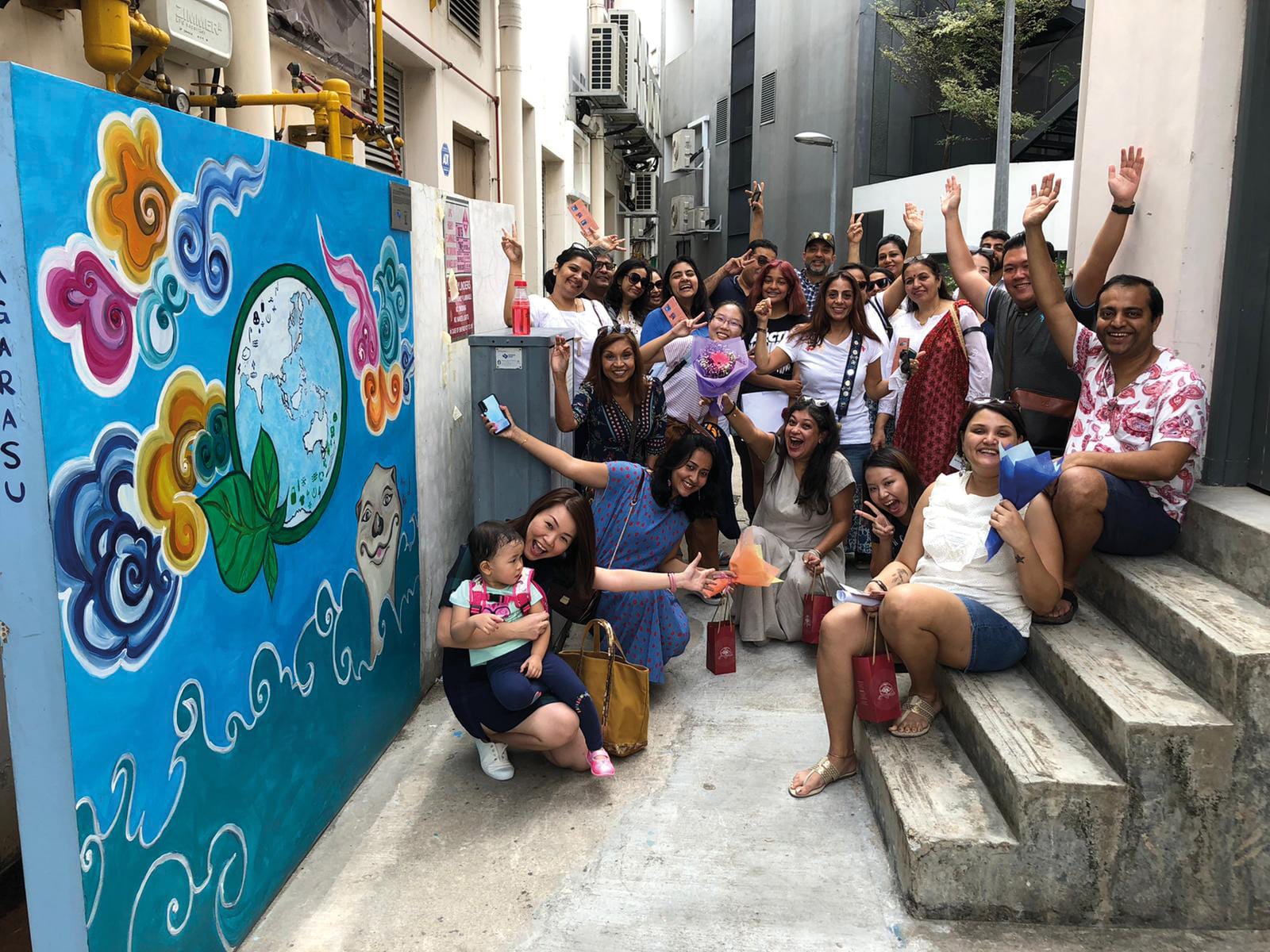 Group of diverse adults and a child, smiling and waving in an alley beside a mural of Earth.